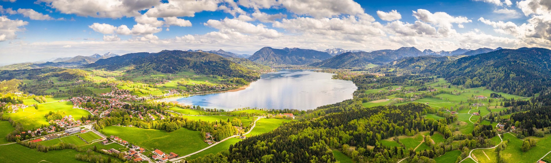 Vogelperspektive des Tegernsees im Sommer inklusive Bergpanorama und umliegender Natur.